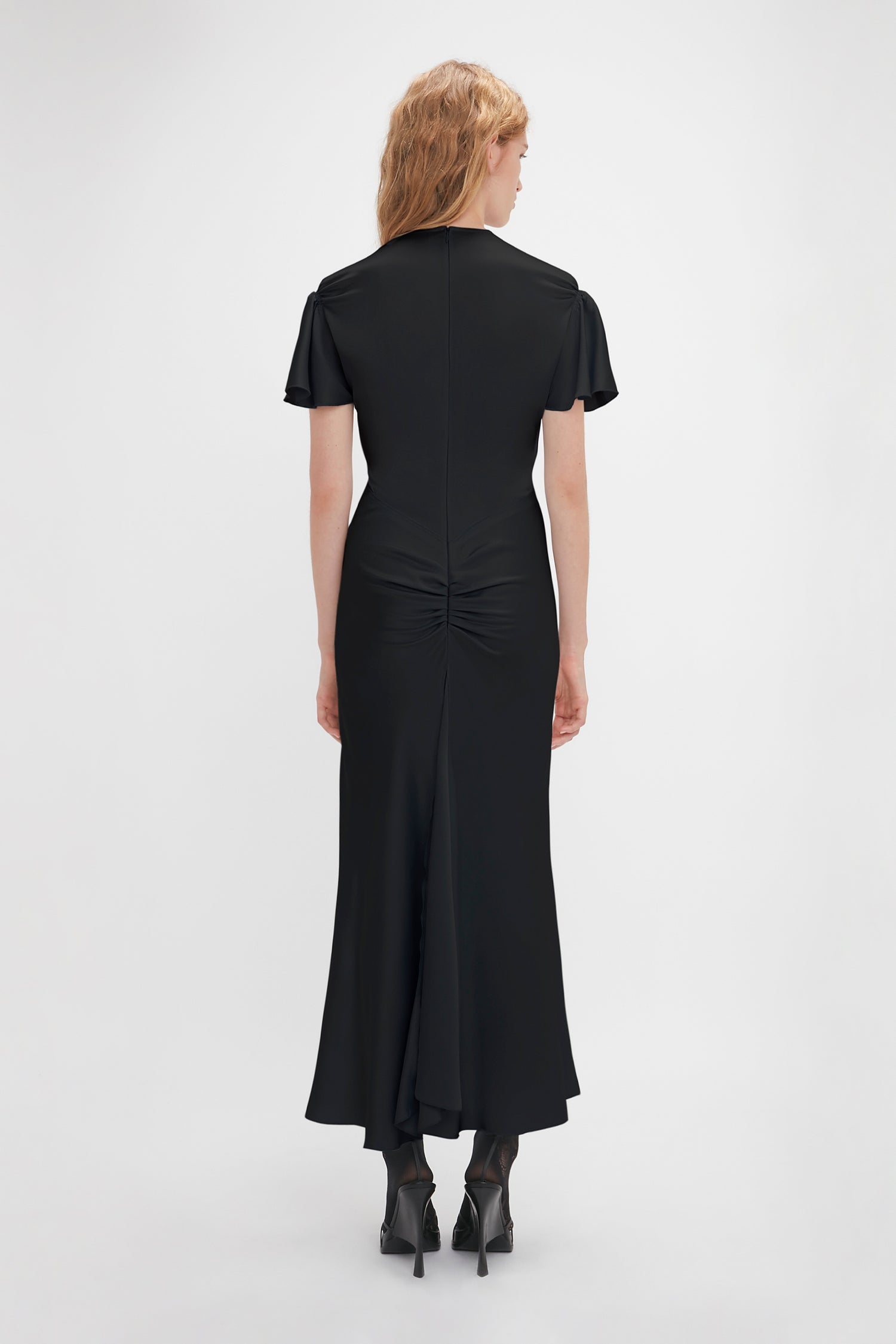 A person with blonde hair stands facing away wearing a Gathered Sleeve Midi Dress In Black by Victoria Beckham, ankle-length, and featuring crepe back satin with ruched detail at the back. They are also wearing black high-heeled shoes.