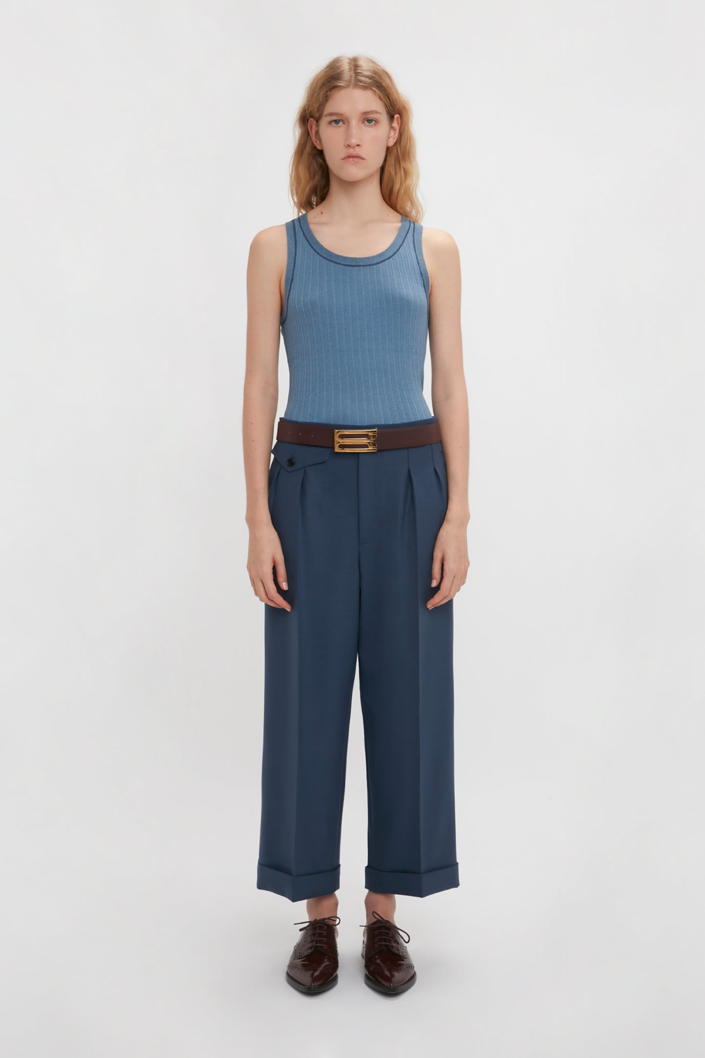 Person standing against a plain background, wearing a Fine Knit Micro Stripe Tank In Heritage Blue by Victoria Beckham, navy high-waisted trousers with a brown belt, and dark brown shoes.