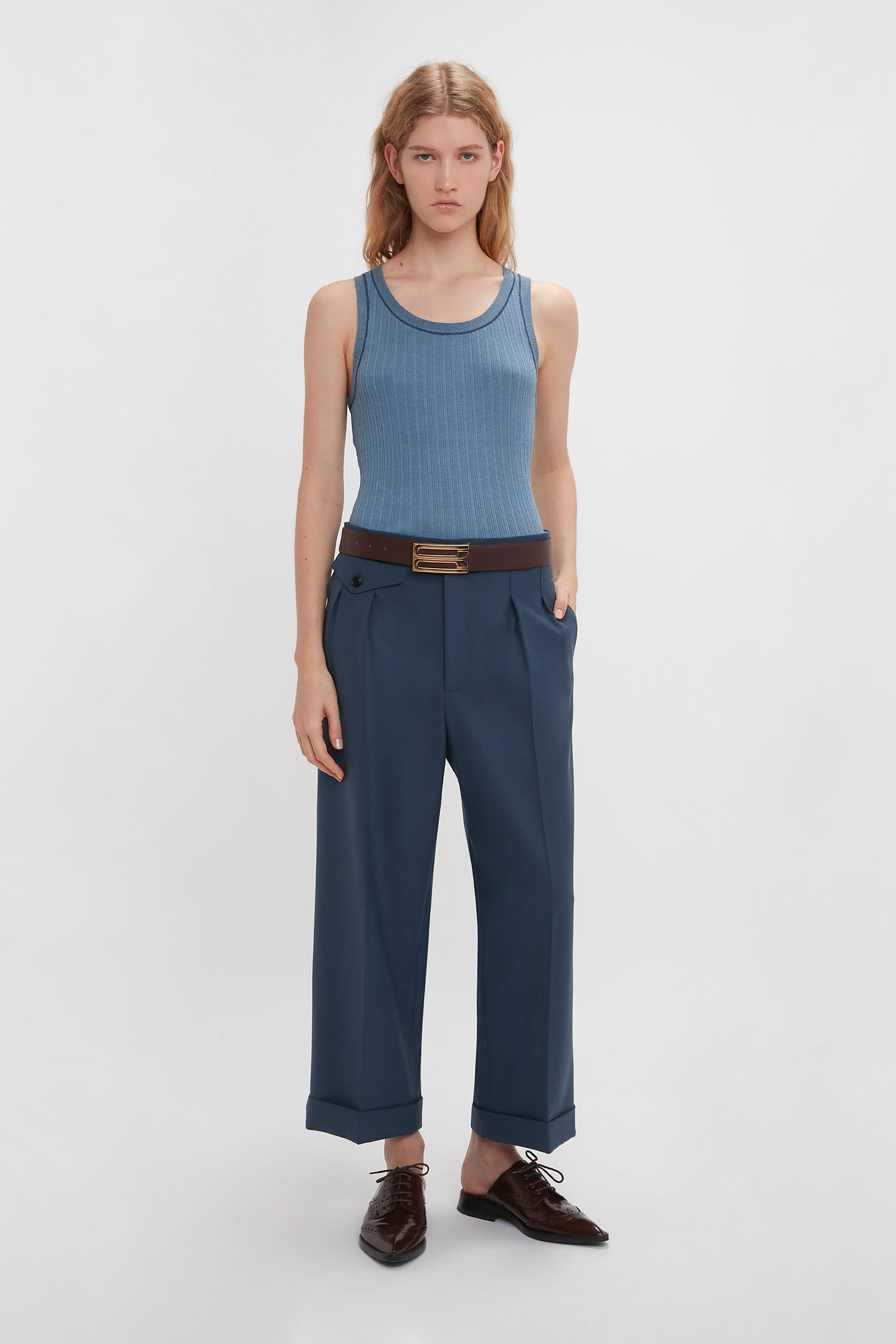 Person standing against a white background, wearing a Victoria Beckham Fine Knit Micro Stripe Tank In Heritage Blue, dark blue trousers with a black belt, and dark brown shoes. Their hands are in their pockets.