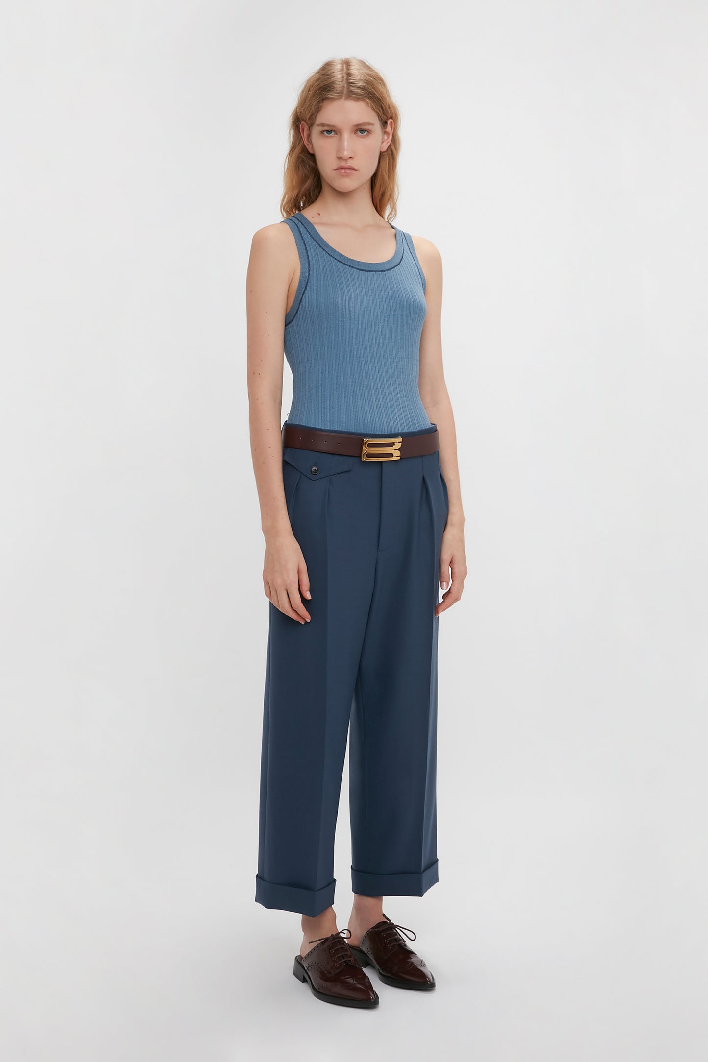 A person with long blonde hair stands against a plain background, wearing a Fine Knit Micro Stripe Tank In Heritage Blue from Victoria Beckham, high-waisted blue pants with a brown belt, and dark brown shoes—reminiscent of Victoria Beckham's timeless style.
