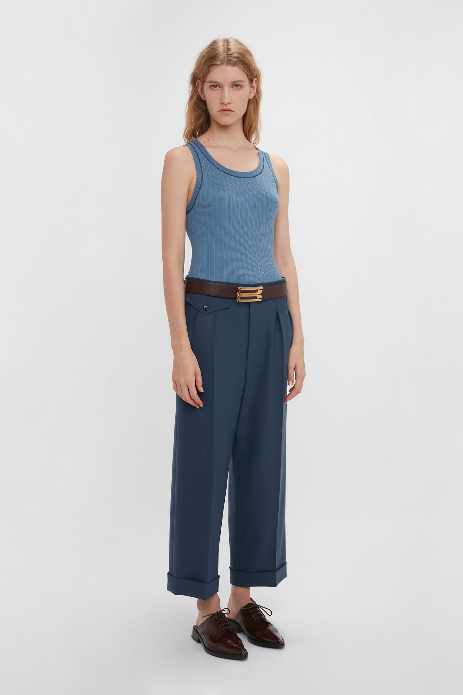 A person with long blonde hair stands against a plain background, wearing a Fine Knit Micro Stripe Tank In Heritage Blue from Victoria Beckham, high-waisted blue pants with a brown belt, and dark brown shoes—reminiscent of Victoria Beckham's timeless style.