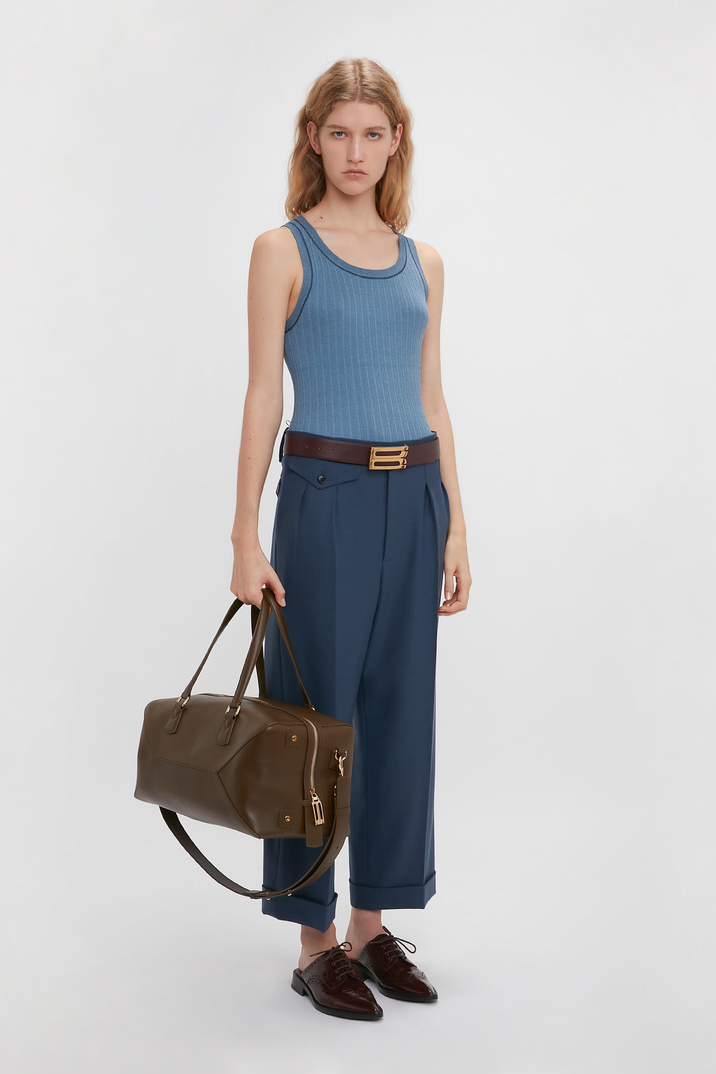 A woman in a blue fine knit tank top and Victoria Beckham navy wide leg cropped trousers with a brown belt, holding a large brown handbag, standing against a white background.