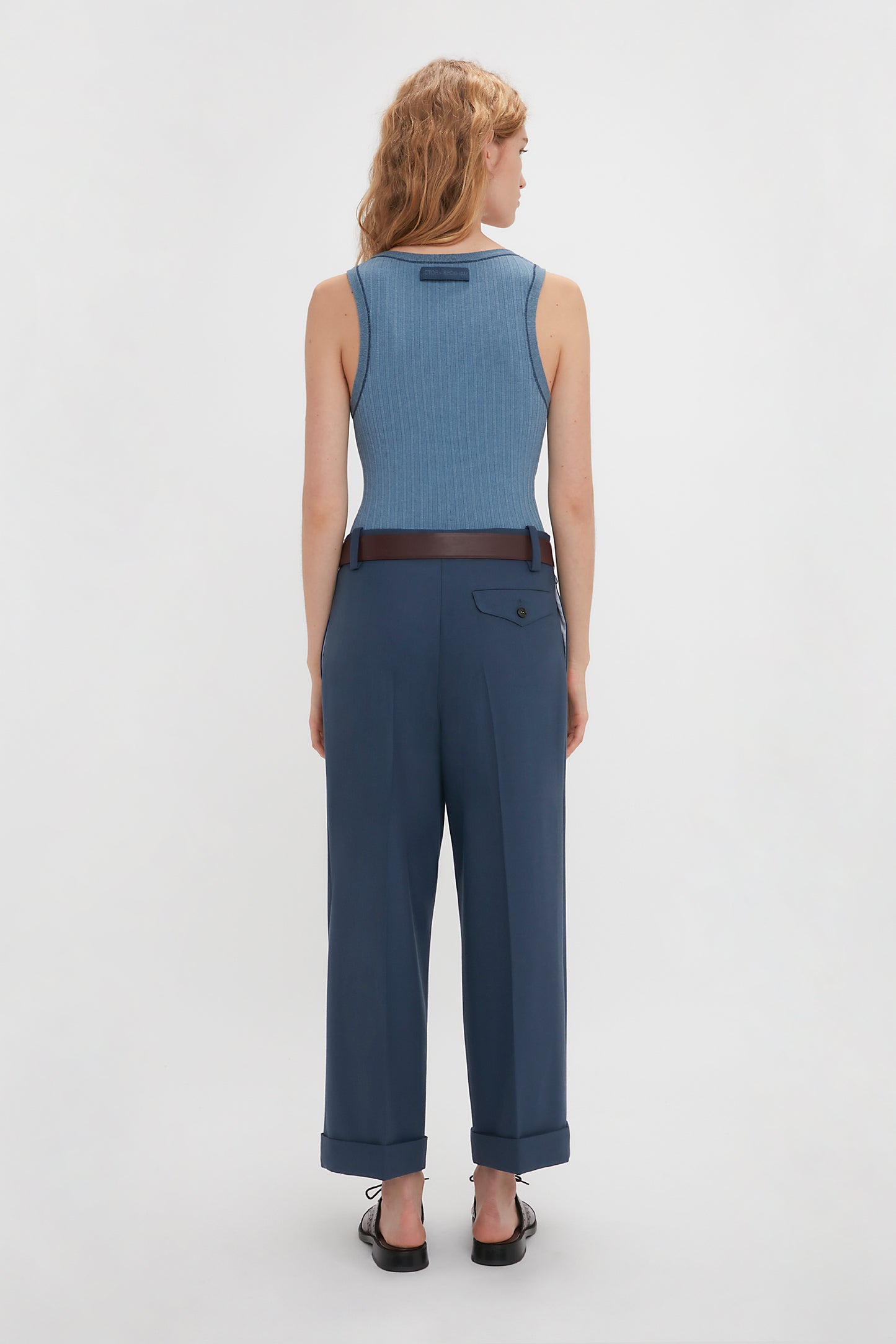 A person with wavy blonde hair is standing with their back to the camera, wearing a Victoria Beckham Fine Knit Micro Stripe Tank In Heritage Blue and dark blue pants, belted at the waist, against a plain white background.