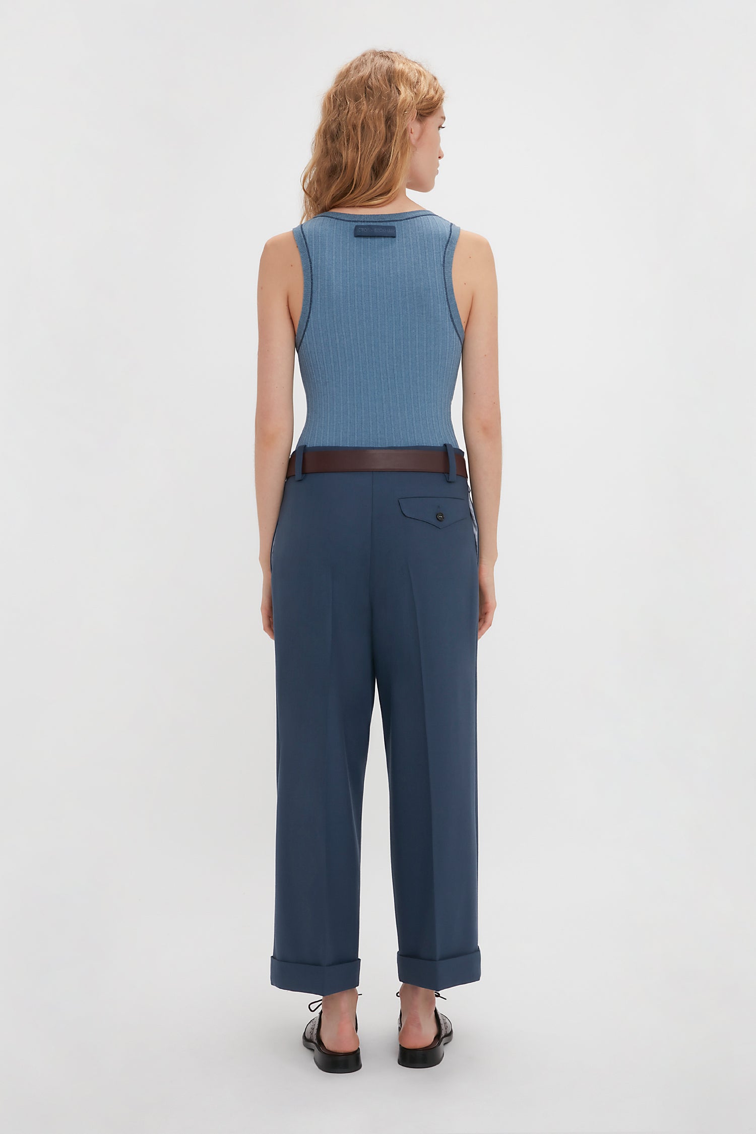 A person with wavy blonde hair is standing with their back to the camera, wearing a Victoria Beckham Fine Knit Micro Stripe Tank In Heritage Blue and dark blue pants, belted at the waist, against a plain white background.