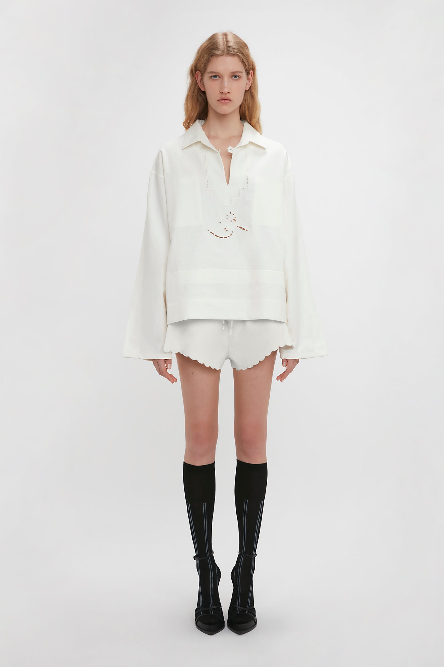 A young woman stands against a white background, wearing the Victoria Beckham Oversized Embroidered Tunic In Antique White and white shorts with scalloped edges, complemented by black knee-high socks with heels.