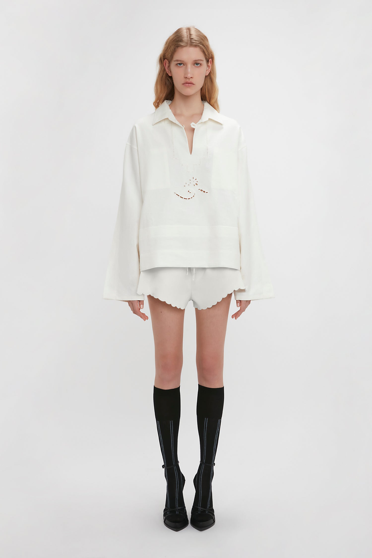 A young woman stands against a white background, wearing the Victoria Beckham Oversized Embroidered Tunic In Antique White and white shorts with scalloped edges, complemented by black knee-high socks with heels.
