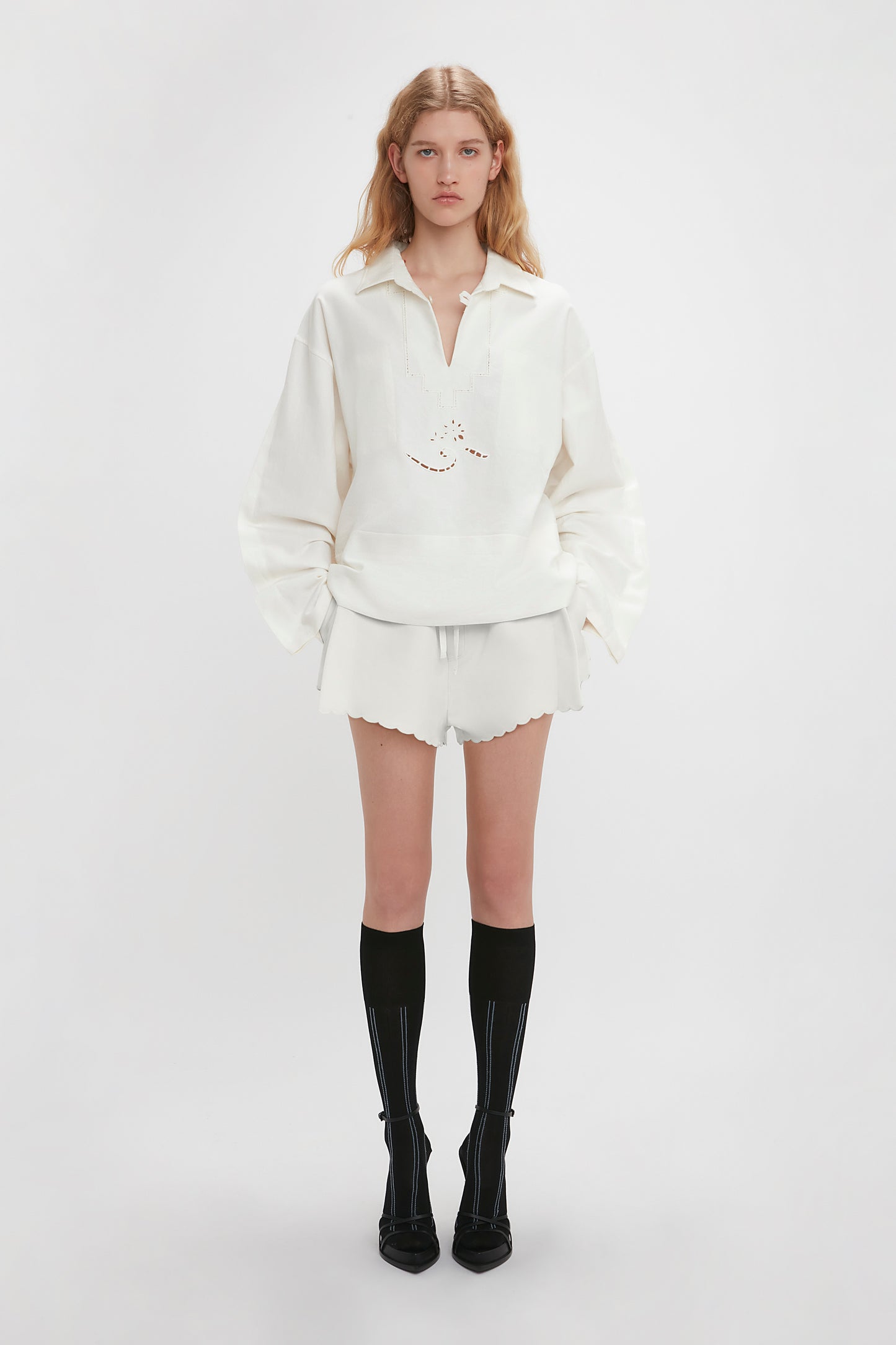 A person stands against a plain background wearing an Antique White pullover, Victoria Beckham Drawstring Embroidered Mini Short In Antique White, and black knee-high socks with black pointed shoes. Their hands are down by their sides.