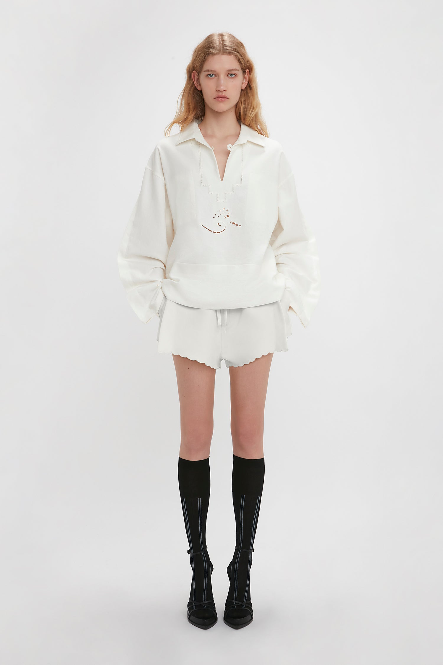 A person stands against a plain background wearing an Antique White pullover, Victoria Beckham Drawstring Embroidered Mini Short In Antique White, and black knee-high socks with black pointed shoes. Their hands are down by their sides.