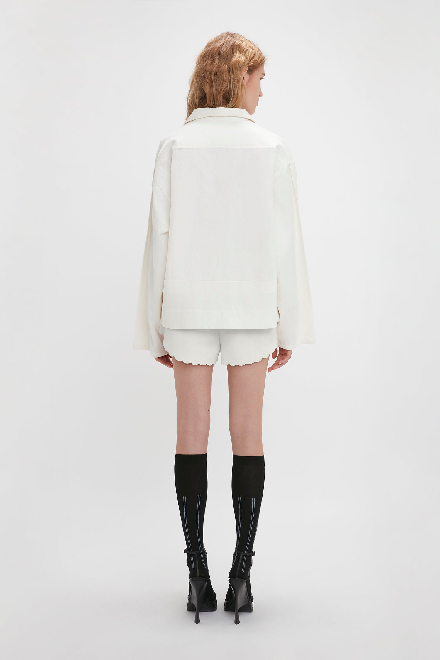 Woman standing with her back to the camera, wearing a white long-sleeve jacket with embroidered detailing, Victoria Beckham Drawstring Embroidered Mini Short In Antique White, black knee-high socks, and black high-heeled shoes against a plain white background.