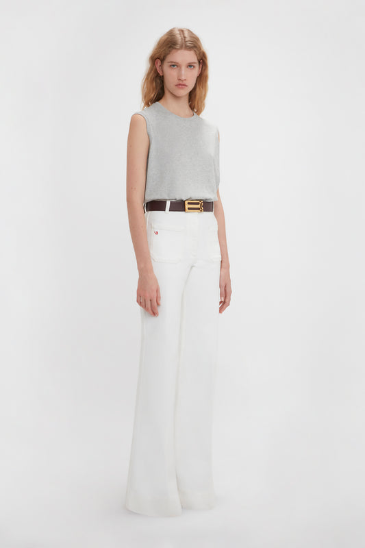 A woman in a Victoria Beckham grey marl sleeveless t-shirt and white wide-legged pants standing against a plain white background. She wears a black belt with a gold buckle.