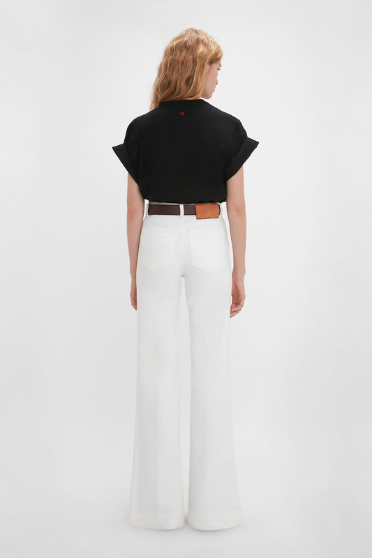 Woman standing with her back to the camera, wearing a black 'Do As I Say, Not As I Do' Slogan T-Shirt by Victoria Beckham and white wide-legged pants with a brown belt.