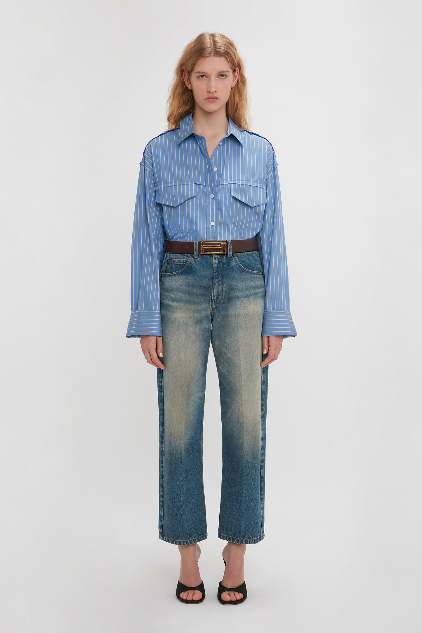 A person stands against a plain background, wearing a Victoria Beckham Cropped Seam Detail Shirt in Steel Blue, faded blue jeans, a black belt, and black heels.