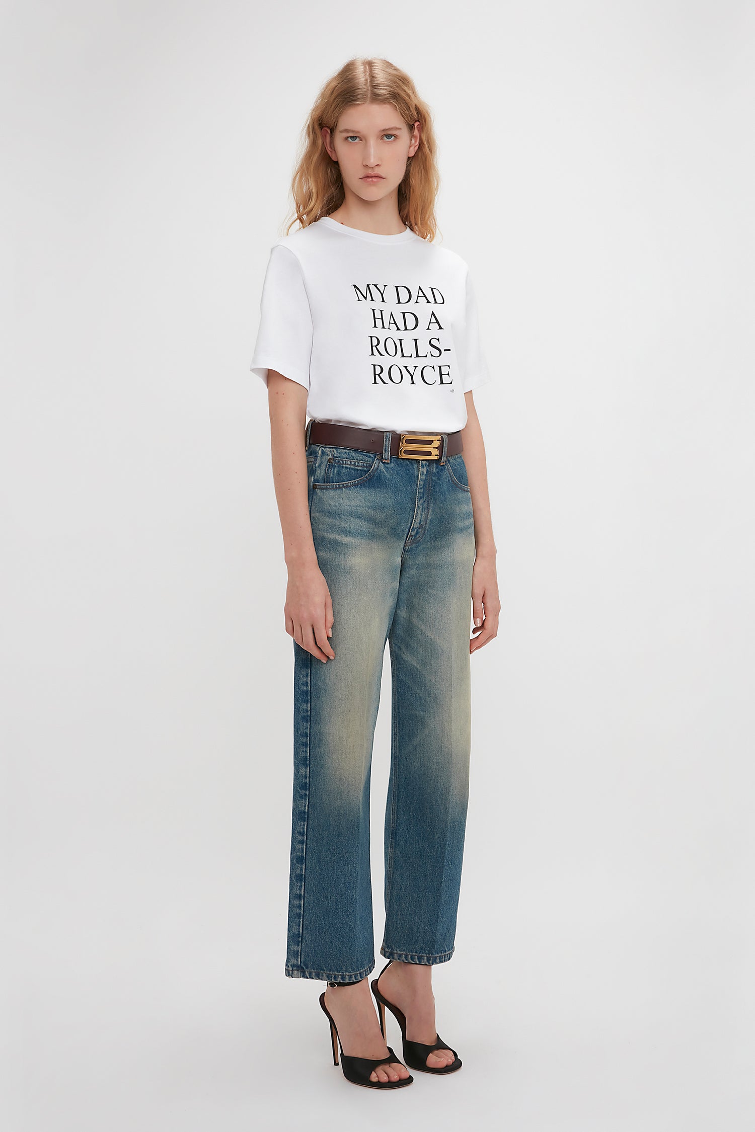 A woman in a white Victoria Beckham Exclusive 'My Dad Had A Rolls-Royce' Slogan T-Shirt and blue jeans, paired with black heels, standing against a white background.