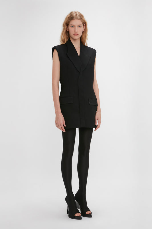 A woman models a Victoria Beckham sleeveless tailored black blazer dress with tights and high-heeled sandals on a plain white background.
