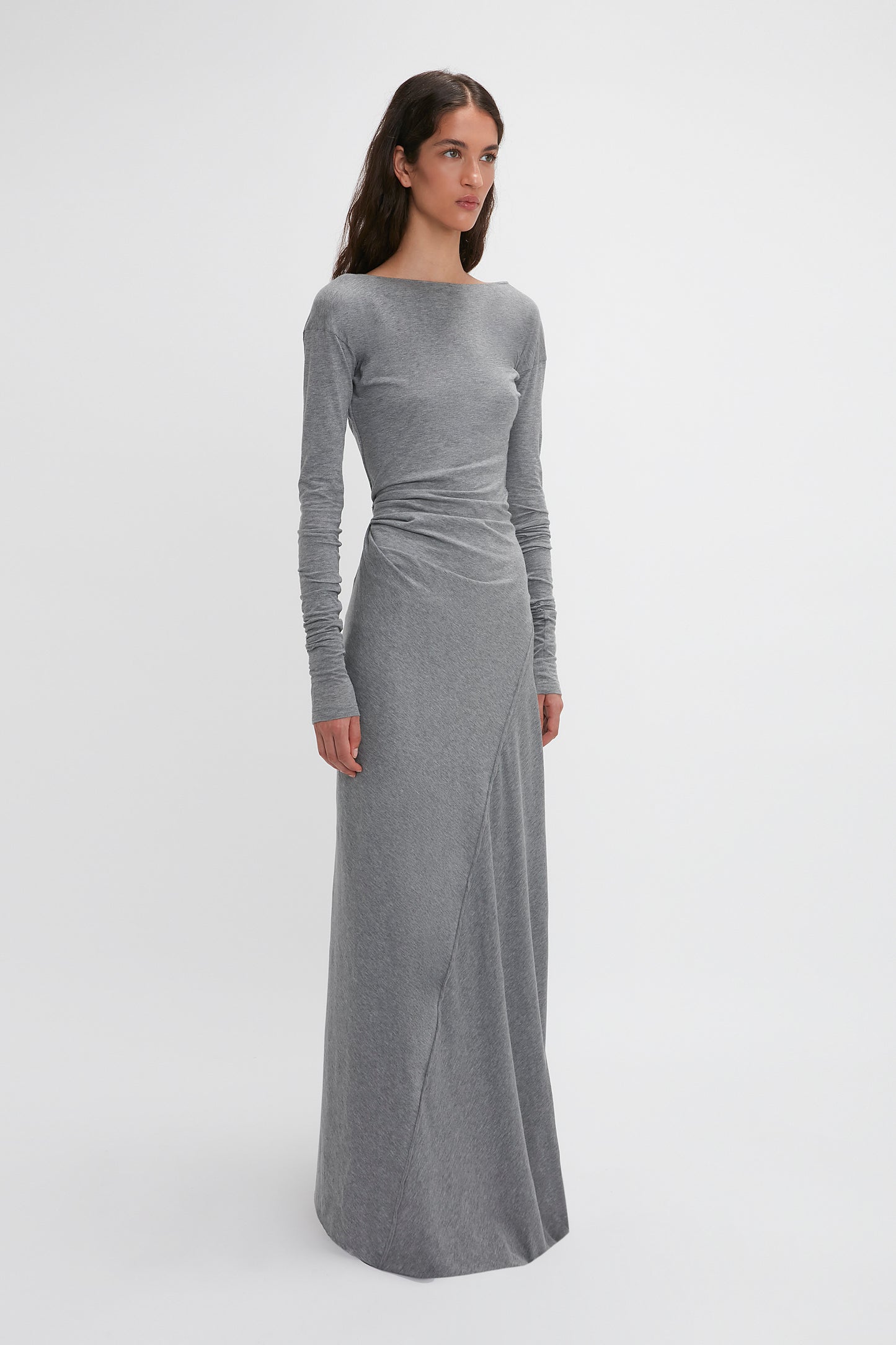 A woman stands against a plain white background wearing the Victoria Beckham Long Sleeve Circle Neck Dress In Grey Marl made from super-soft cotton-jersey blend that reaches the floor. She looks slightly to the side with a neutral expression, epitomizing contemporary eveningwear.