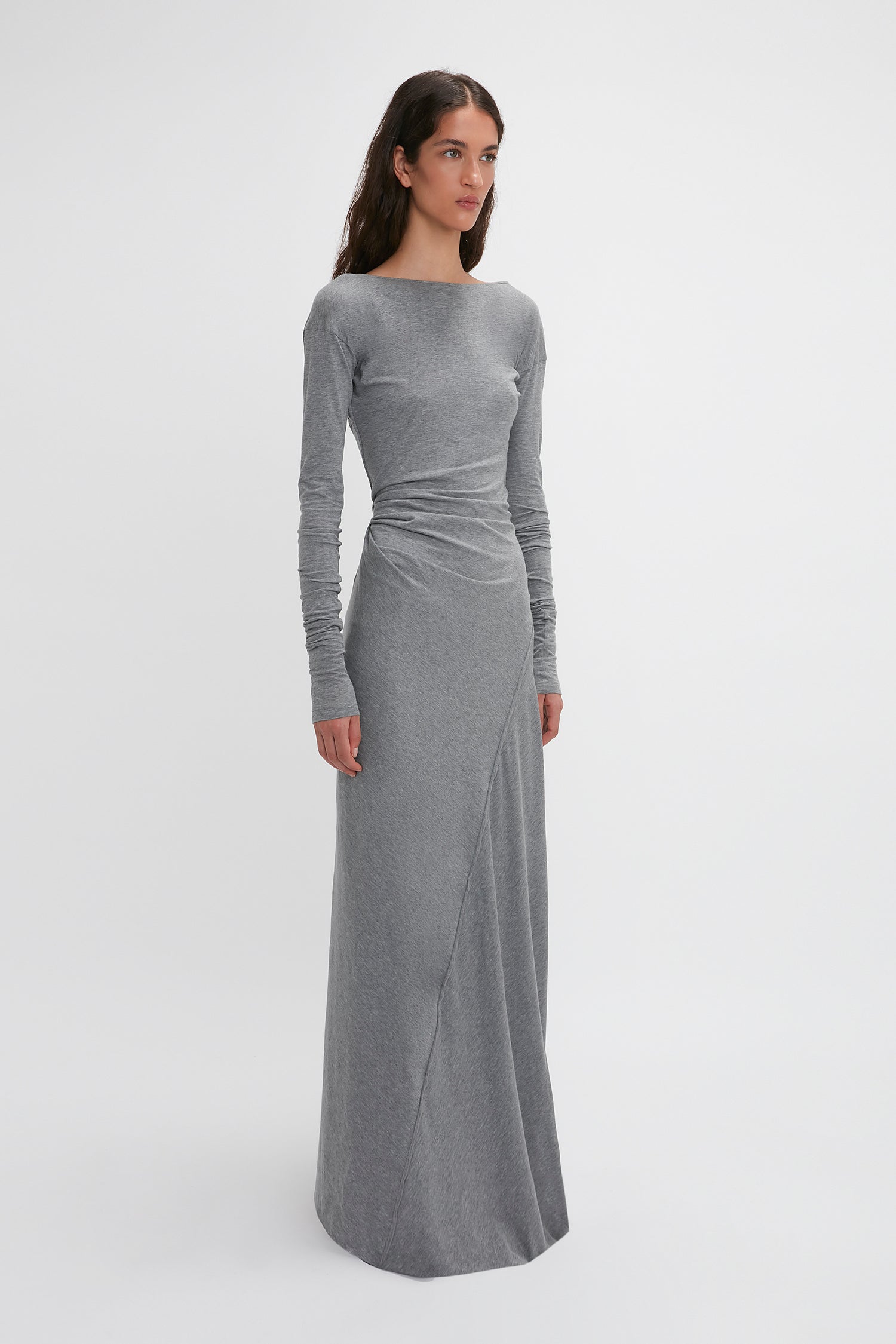 A woman stands against a plain white background wearing the Victoria Beckham Long Sleeve Circle Neck Dress In Grey Marl made from super-soft cotton-jersey blend that reaches the floor. She looks slightly to the side with a neutral expression, epitomizing contemporary eveningwear.