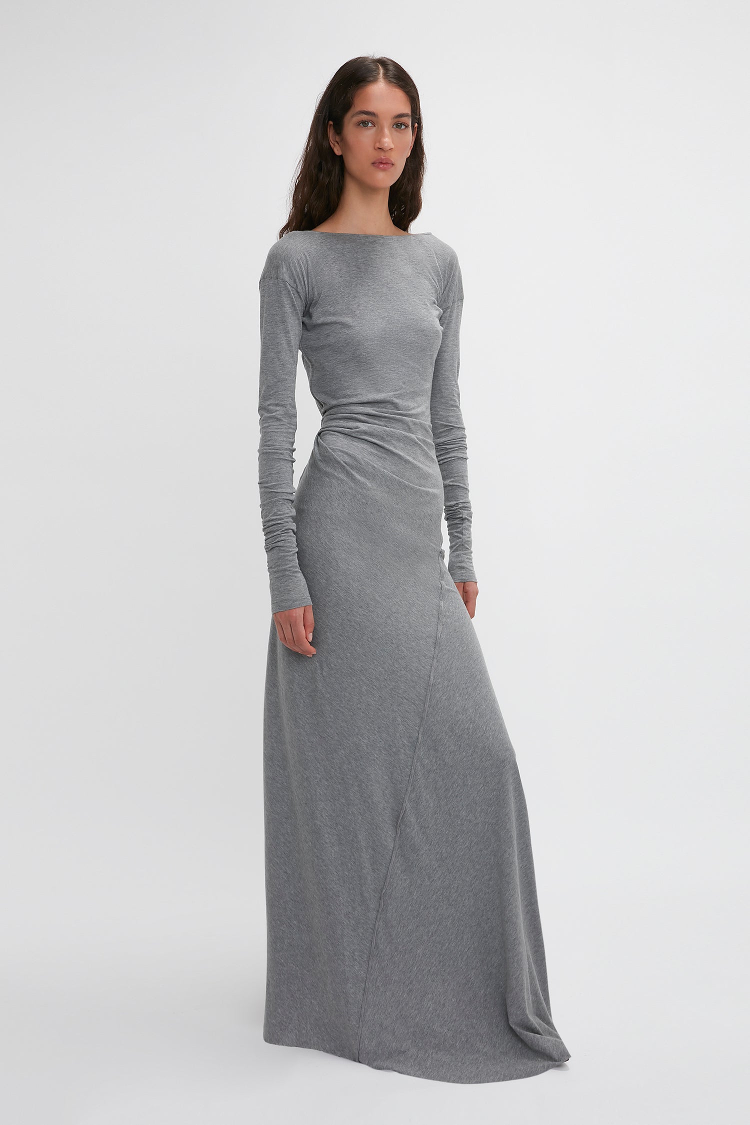 A woman in a long-sleeved, floor-length gray Long Sleeve Circle Neck Dress In Grey Marl from Victoria Beckham, made from a super-soft cotton-jersey blend, stands posing against a plain white background.