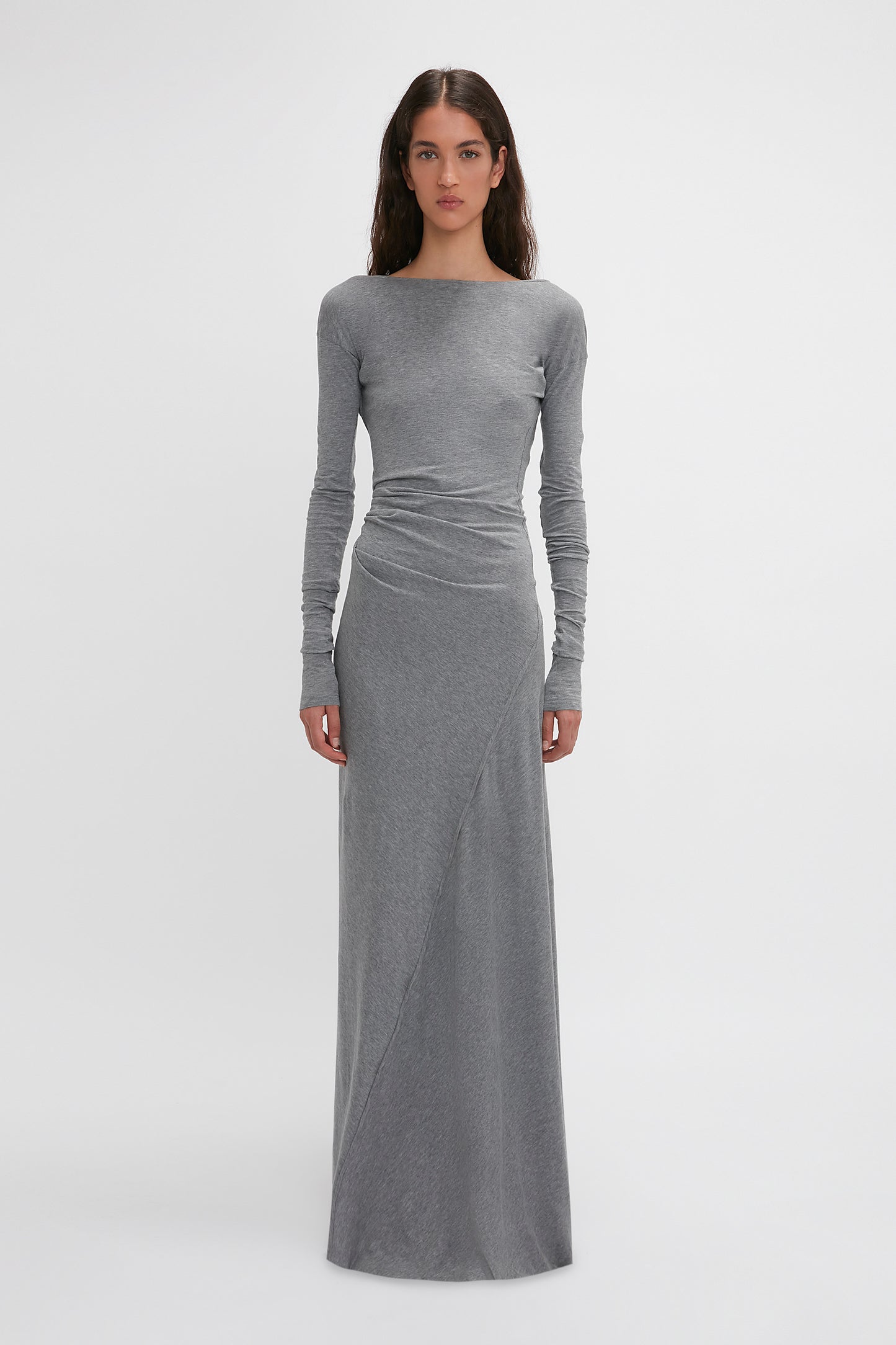 A woman stands facing forward wearing the Long Sleeve Circle Neck Dress In Grey Marl by Victoria Beckham, a long-sleeved, fitted gray maxi dress with a slight draping effect at the waist, made from a super-soft cotton-jersey blend, set against a plain white background.