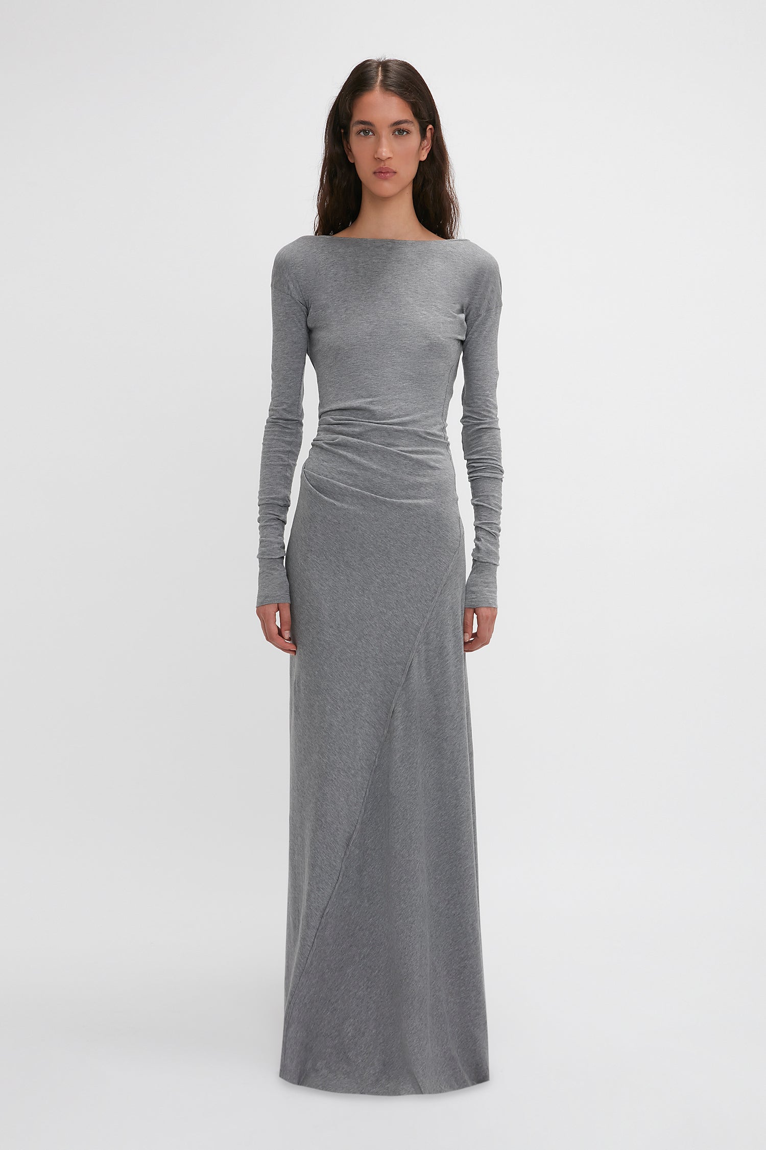 A woman stands facing forward wearing the Long Sleeve Circle Neck Dress In Grey Marl by Victoria Beckham, a long-sleeved, fitted gray maxi dress with a slight draping effect at the waist, made from a super-soft cotton-jersey blend, set against a plain white background.