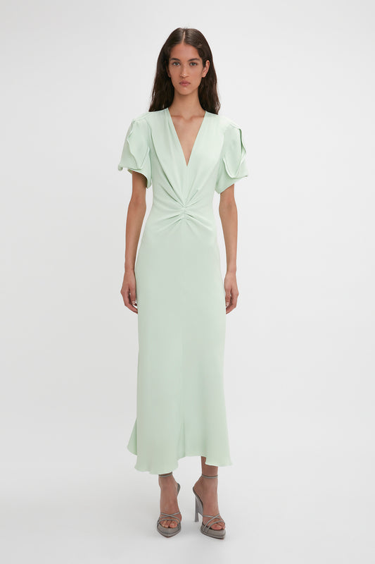 A woman stands against a plain background, wearing a light green, Gathered V-neck Midi Dress In Jade with short sleeves, waist-defining pleat detail, and silver heels by Victoria Beckham.