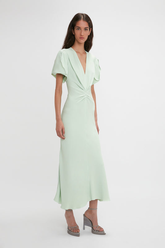 A woman stands against a white background, wearing a Victoria Beckham light green Gathered V-Neck Midi Dress In Jade with short, puffy sleeves and waist-defining pleat detail. She is also wearing silver high-heeled sandals.