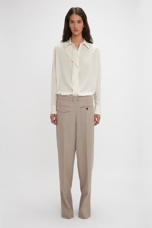A person stands against a plain background wearing an Asymmetric Ruffle Blouse In Ivory by Victoria Beckham, featuring voluminous sleeves, and beige trousers.
