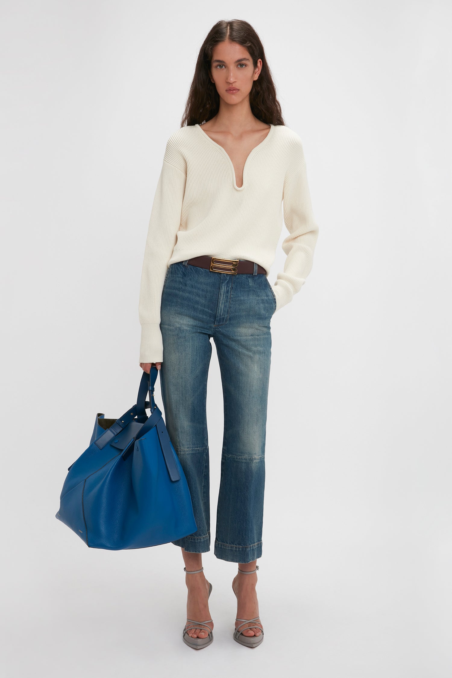 A woman stands against a plain background wearing a white sweater, blue jeans, and heels, while holding a Victoria Beckham W11 Medium Tote Bag In Vibrant Blue Leather.