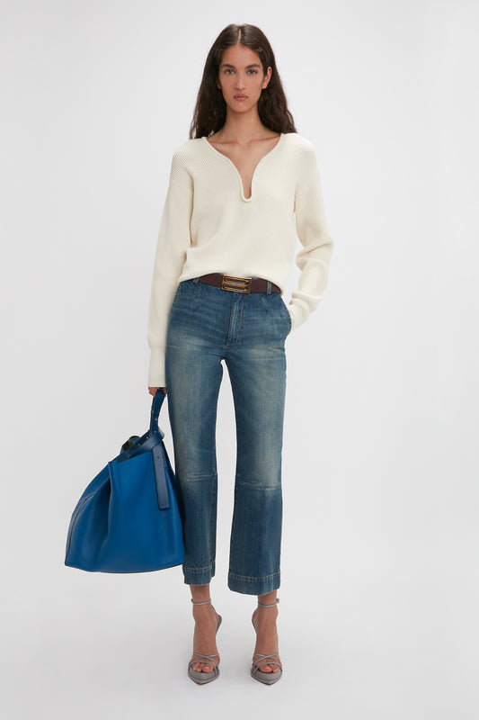 A woman stands in a studio, wearing a white sweater, Victoria Beckham cropped kick jeans in Indigrey wash, and sandals, holding a blue tote bag.