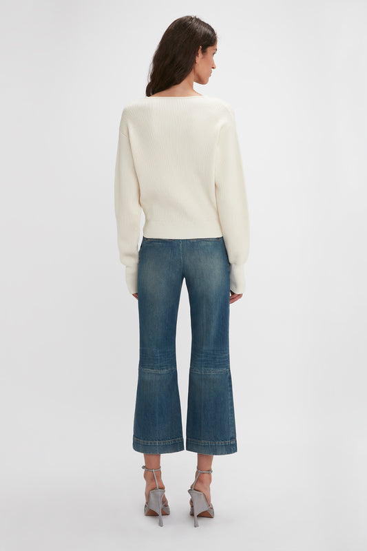 Woman wearing a cream sweater and Victoria Beckham Cropped Kick Jean In Indigrey Wash with a flared silhouette, viewed from the back, standing against a white background.