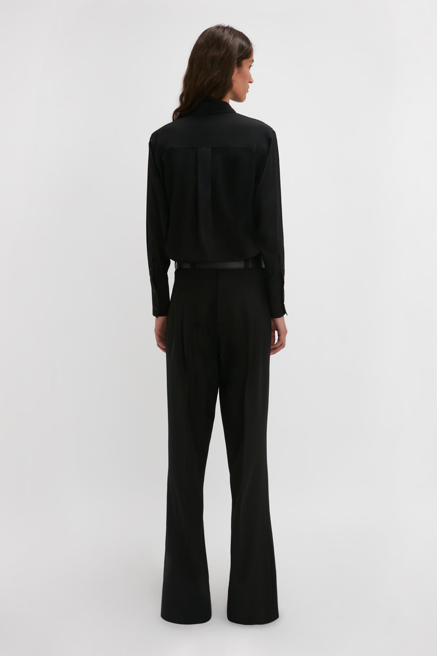 A woman in a black pantsuit stands facing away, showcasing the tailored back design of the Victoria Beckham Reverse Front Trouser In Black on a plain light background, highlighting the contemporary silhouette.