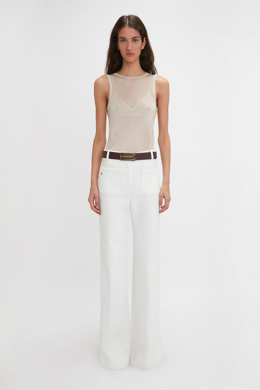 A person stands against a plain white background, wearing the Victoria Beckham Lightweight Tank Top In Birch, paired with high-waisted, wide-leg white pants and a dark belt.