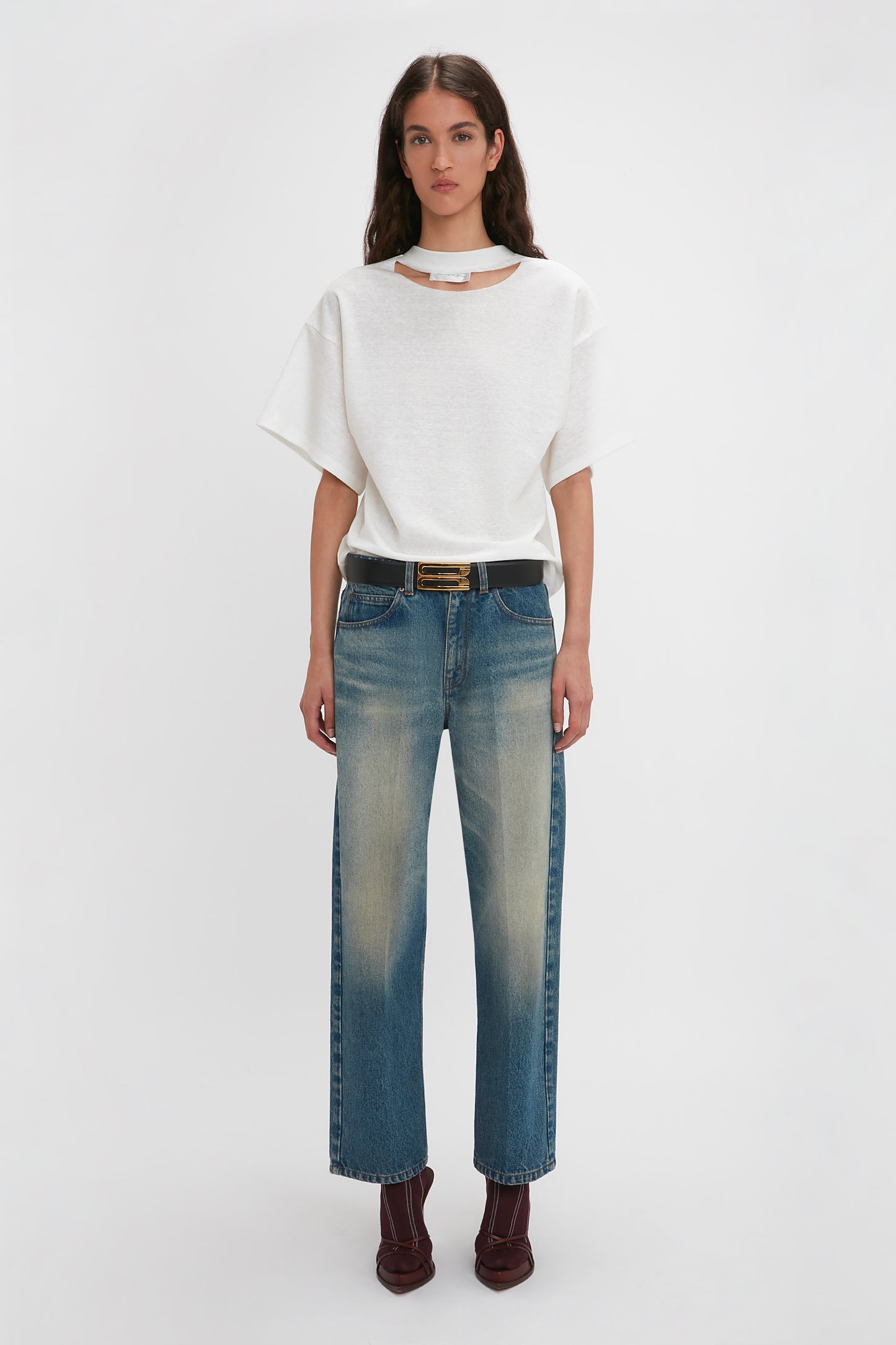 A person stands against a plain white background, wearing a relaxed cut Victoria Beckham Bonded Jersey Tee In White, blue jeans with light wash details, a black belt, and brown ankle boots.