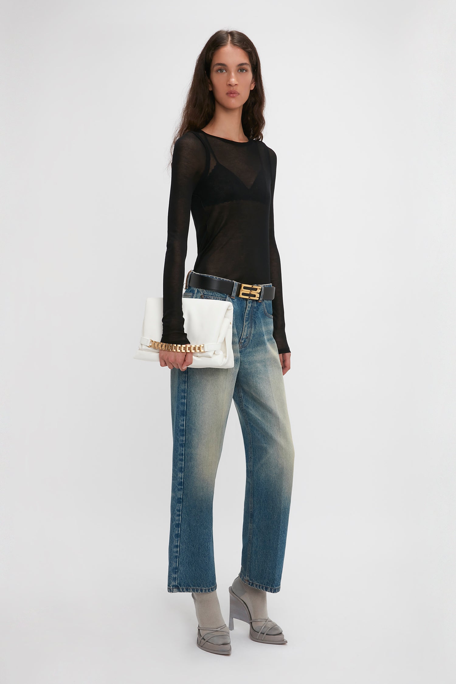 A woman in a black Crew Neck Long Sleeve Top In Black by Victoria Beckham, blue jeans, and gray high heels holds a white clutch bag against a plain background. The top, made of semi-sheer fabric, adds an elegant touch to her ensemble.