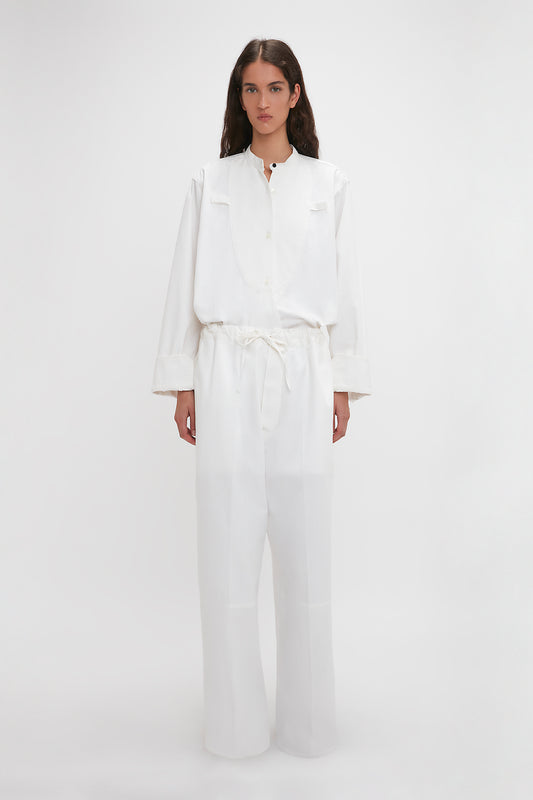 A woman stands against a white background wearing a stylish white blouse and matching cotton-canvas Victoria Beckham Drawstring Pyjama Trousers in Washed White.