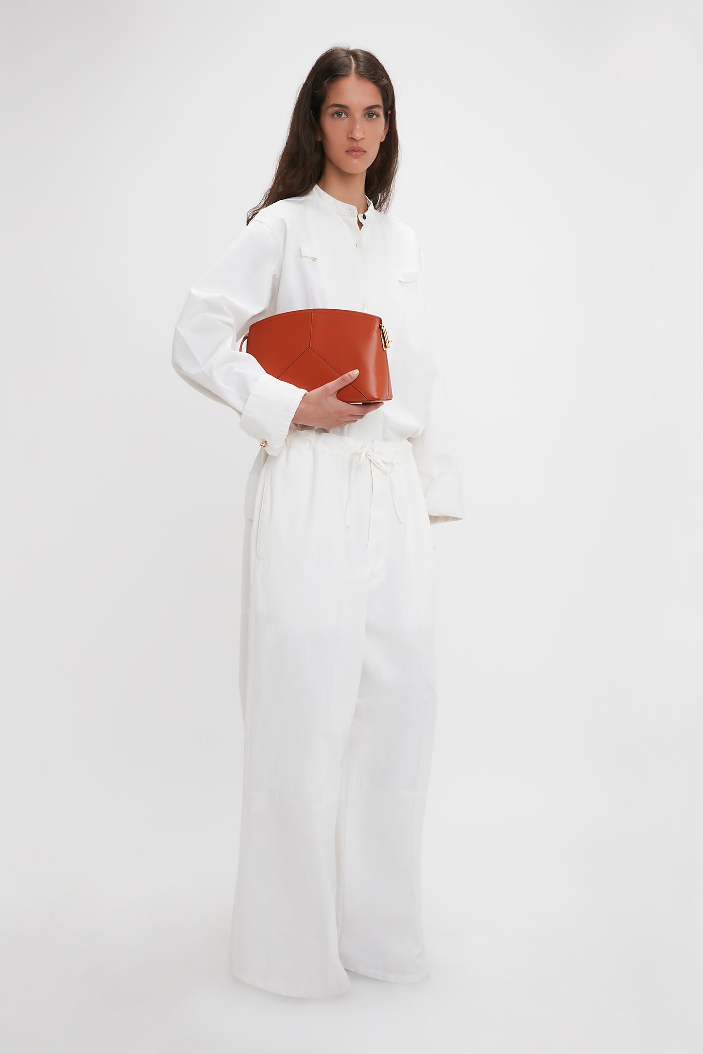 Woman in a white outfit holding a brown geometric Victoria Beckham Victoria Clutch Bag In Tan Leather made of glossy calf leather with a branded padlock, standing against a plain white background.