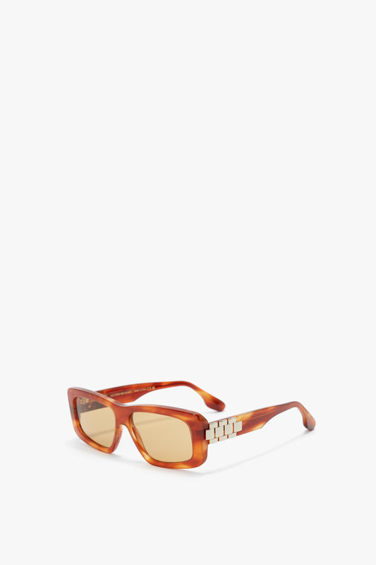Rectangular amber-colored sunglasses with tinted lenses and metal decorative accents on the temples, embodying the sophisticated style of Victoria Beckham eyewear. The Chain Detail Rectangular Frame Sunglasses In Striped Blonde Havana is elegantly showcased against a pristine white background.