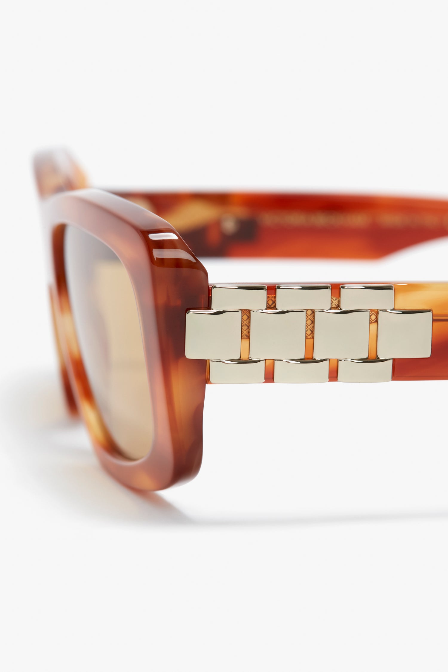 Close-up of tortoiseshell Victoria Beckham Chain Detail Rectangular Frame Sunglasses In Striped Blonde Havana with gold chain detail on the temple.