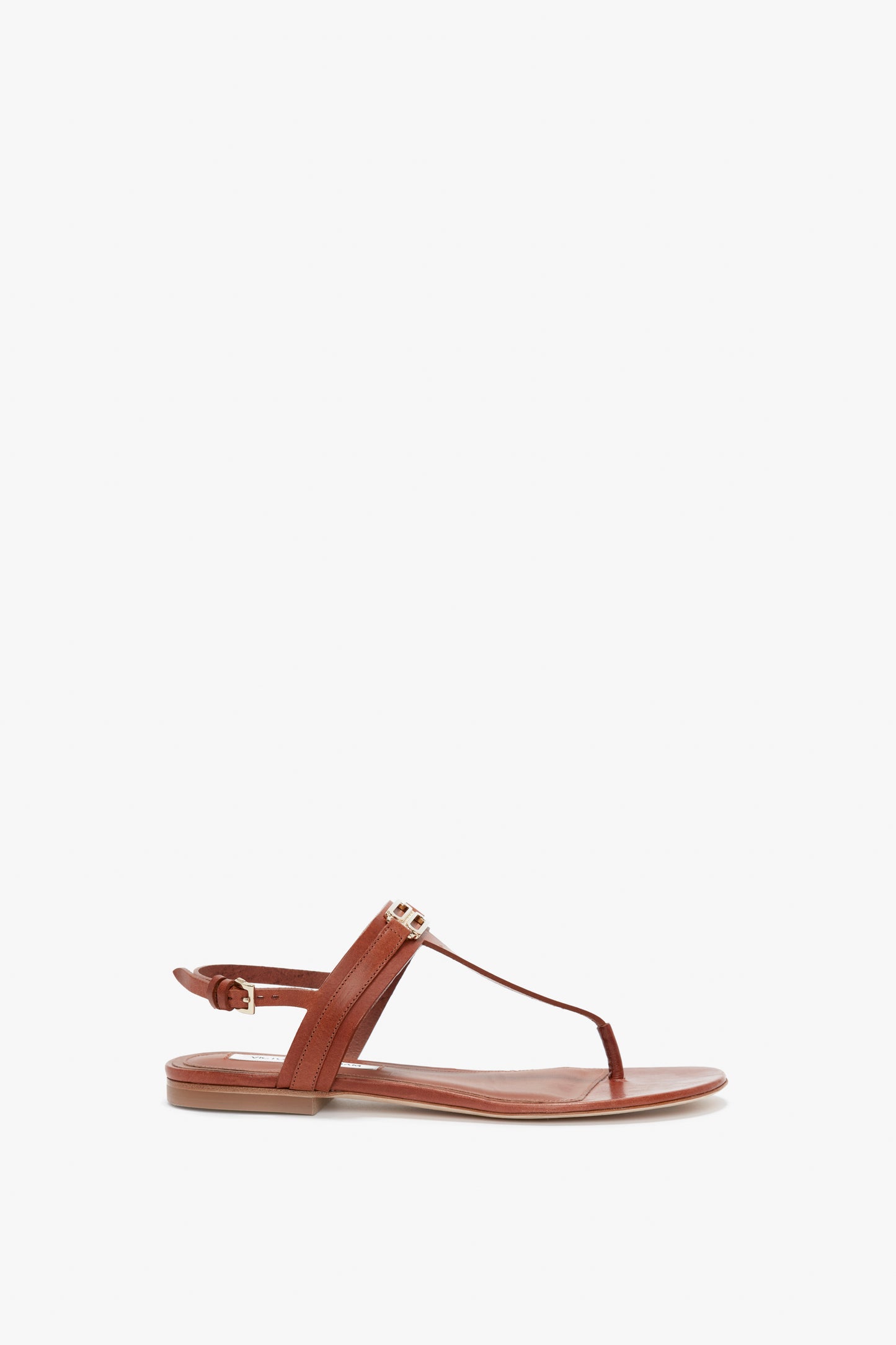 A single brown flat sandal with a thong-style strap, an adjustable ankle strap, and a simple, minimalist design featuring a subtle gold-tone chain detail by Victoria Beckham is the Flat Chain Sandal in Tan Leather.