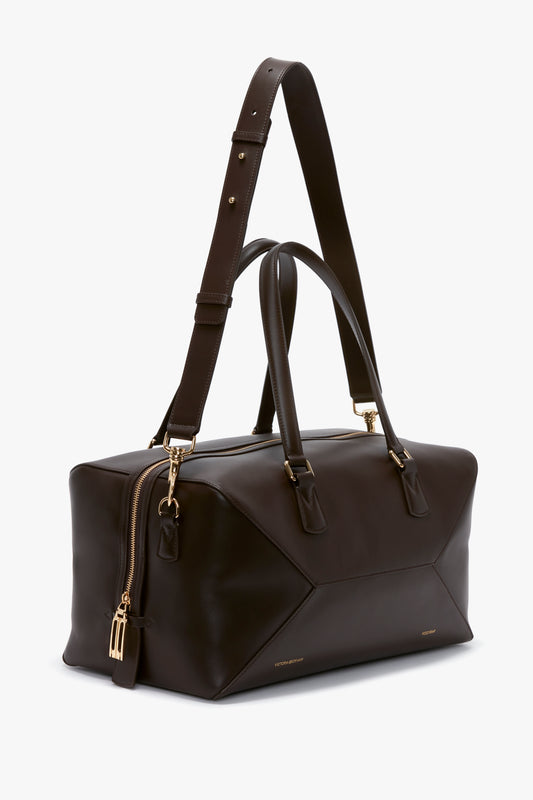 A dark brown leather duffel bag made of luxe calfskin with gold zippers and hardware. It features two short handles and a long, adjustable shoulder strap. This Medium Gym Bag In Chocolate Brown by Victoria Beckham exudes a timeless elegance reminiscent of Victoria Beckham's sophisticated style.