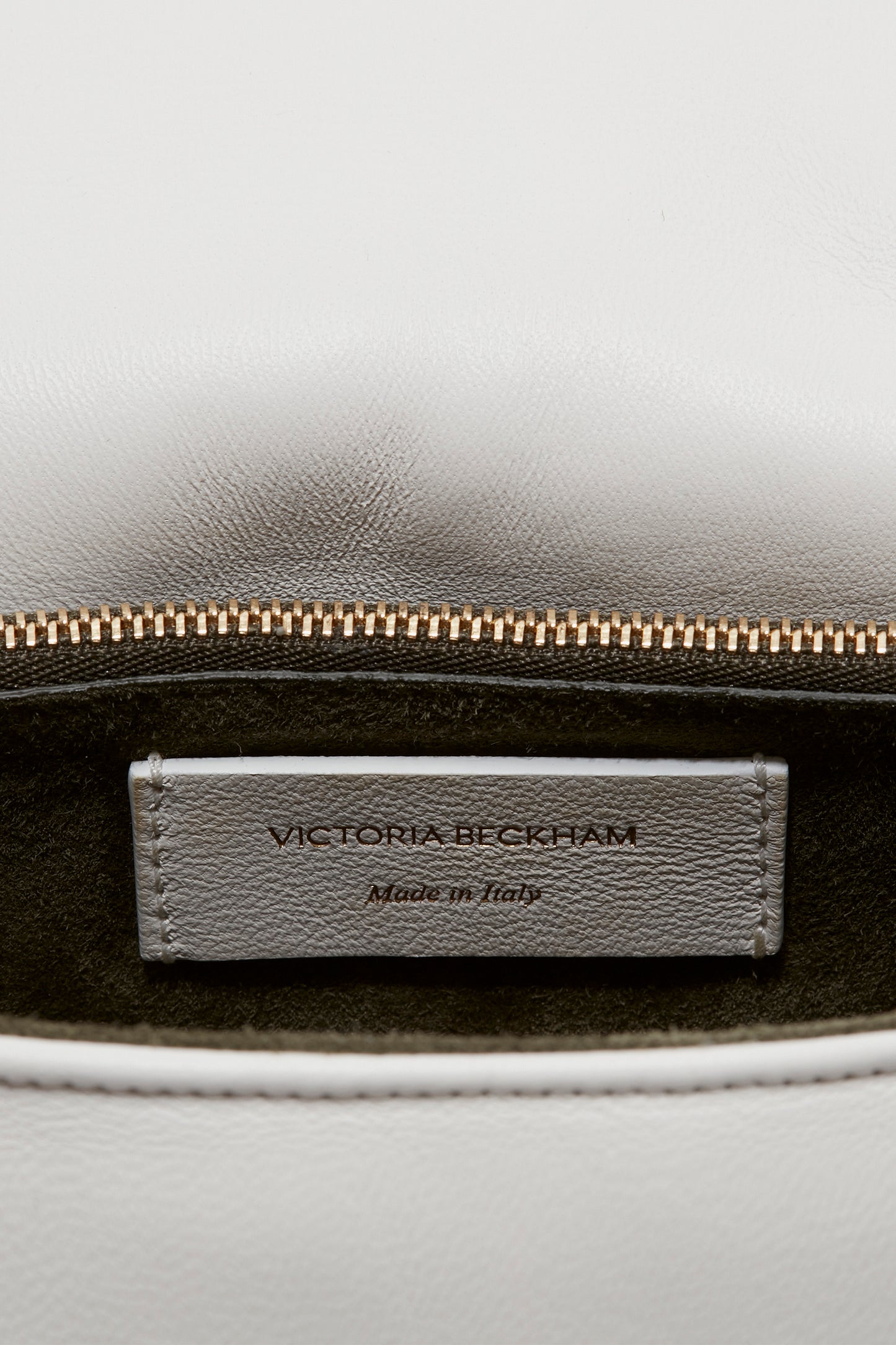 Close-up of a Victoria Beckham Puffy Chain Pouch With Strap In White Leather's interior label, showing "VICTORIA BECKHAM" and "Made in Italy" stitched on a gray leather tag, with a partially open gold zipper and luxurious gold chain detail above it.
