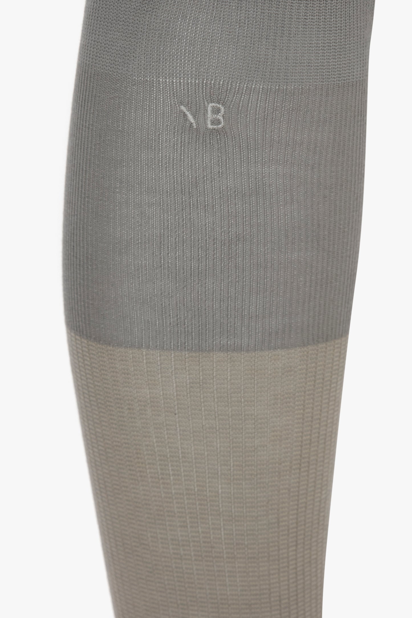 A close-up image of a grey and beige compression sock, showing a gradient transition between the two colors and a faintly embossed "B" near the top. These Superfine Rib Socks In Lunar Grey by Victoria Beckham are an elevated wardrobe essential designed with meticulous detail in 100% cotton.