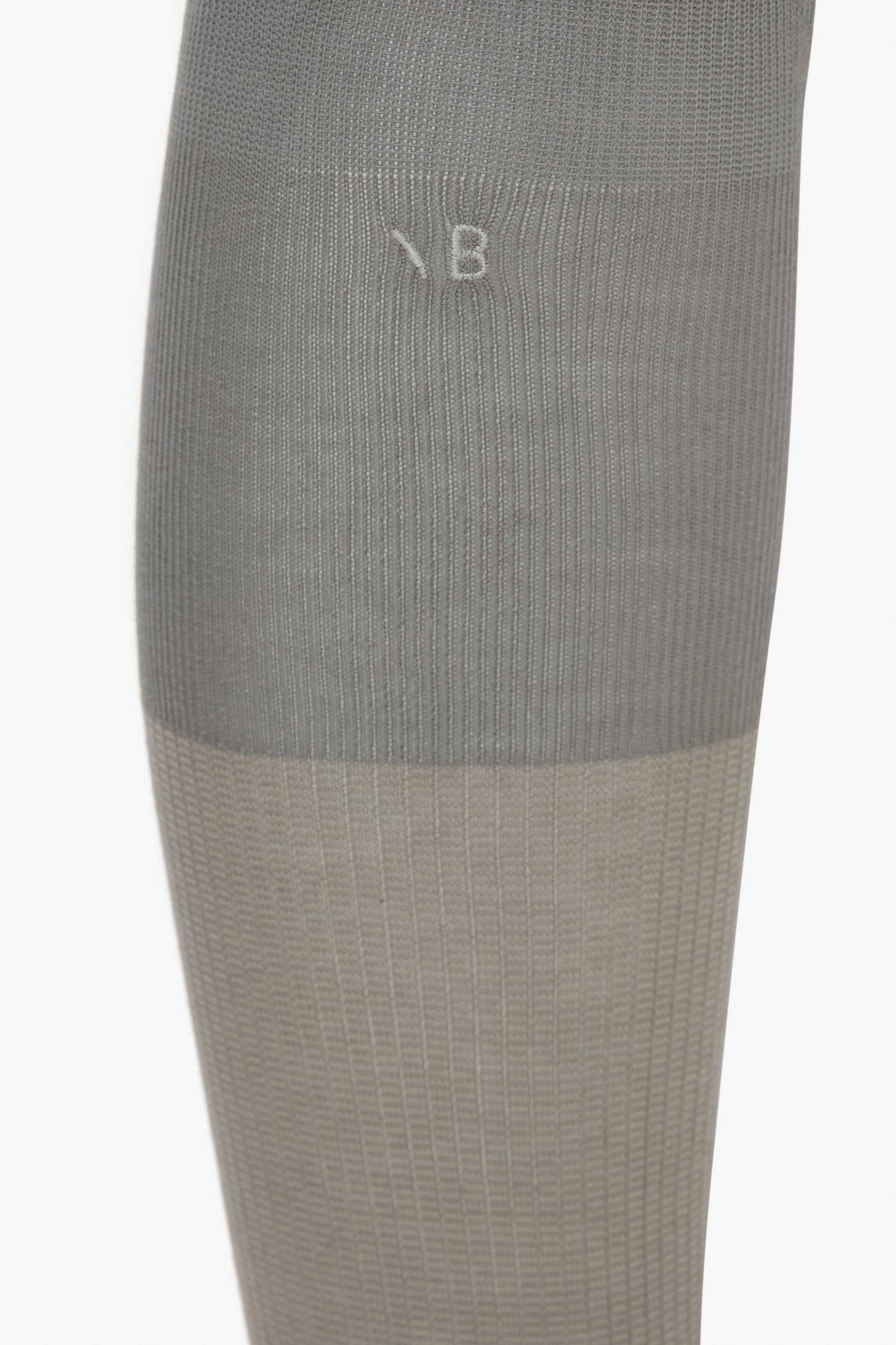 A close-up image of a grey and beige compression sock, showing a gradient transition between the two colors and a faintly embossed "B" near the top. These Superfine Rib Socks In Lunar Grey by Victoria Beckham are an elevated wardrobe essential designed with meticulous detail in 100% cotton.
