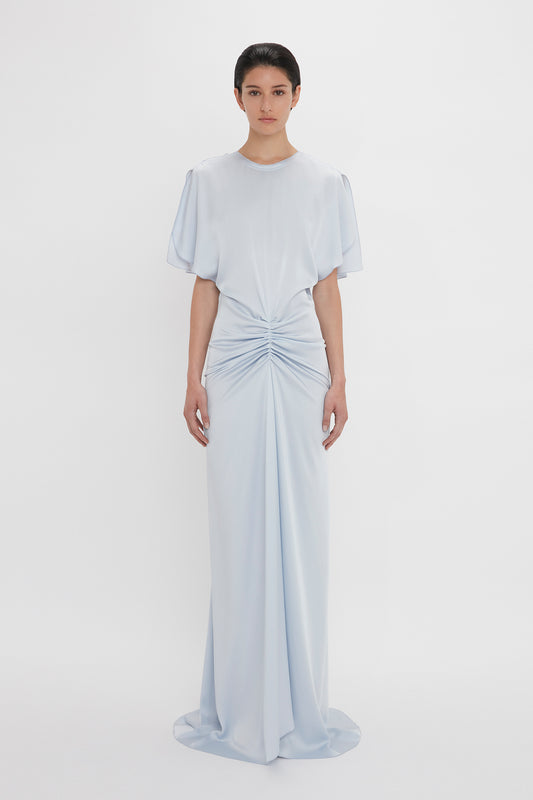 A person wearing the Exclusive Floor-Length Gathered Dress In Ice by Victoria Beckham stands against a plain white background. The gown elegantly features an open back and a waist-defining silhouette that flatters the figure perfectly.