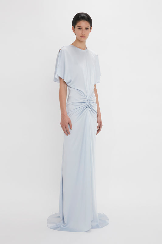 A person stands wearing the Exclusive Floor-Length Gathered Dress In Ice by Victoria Beckham, a light blue, short-sleeved, floor-length dress with gathered detailing at the waist, creating a waist-defining silhouette against a plain white background.