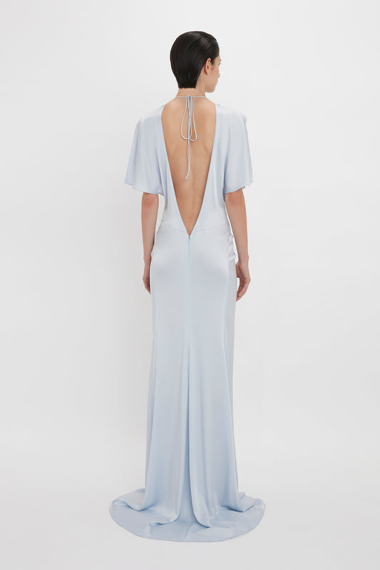 Person wearing the Victoria Beckham Exclusive Floor-Length Gathered Dress In Ice with an open back, showcasing a waist-defining silhouette, stands facing away from the camera against a plain white background.