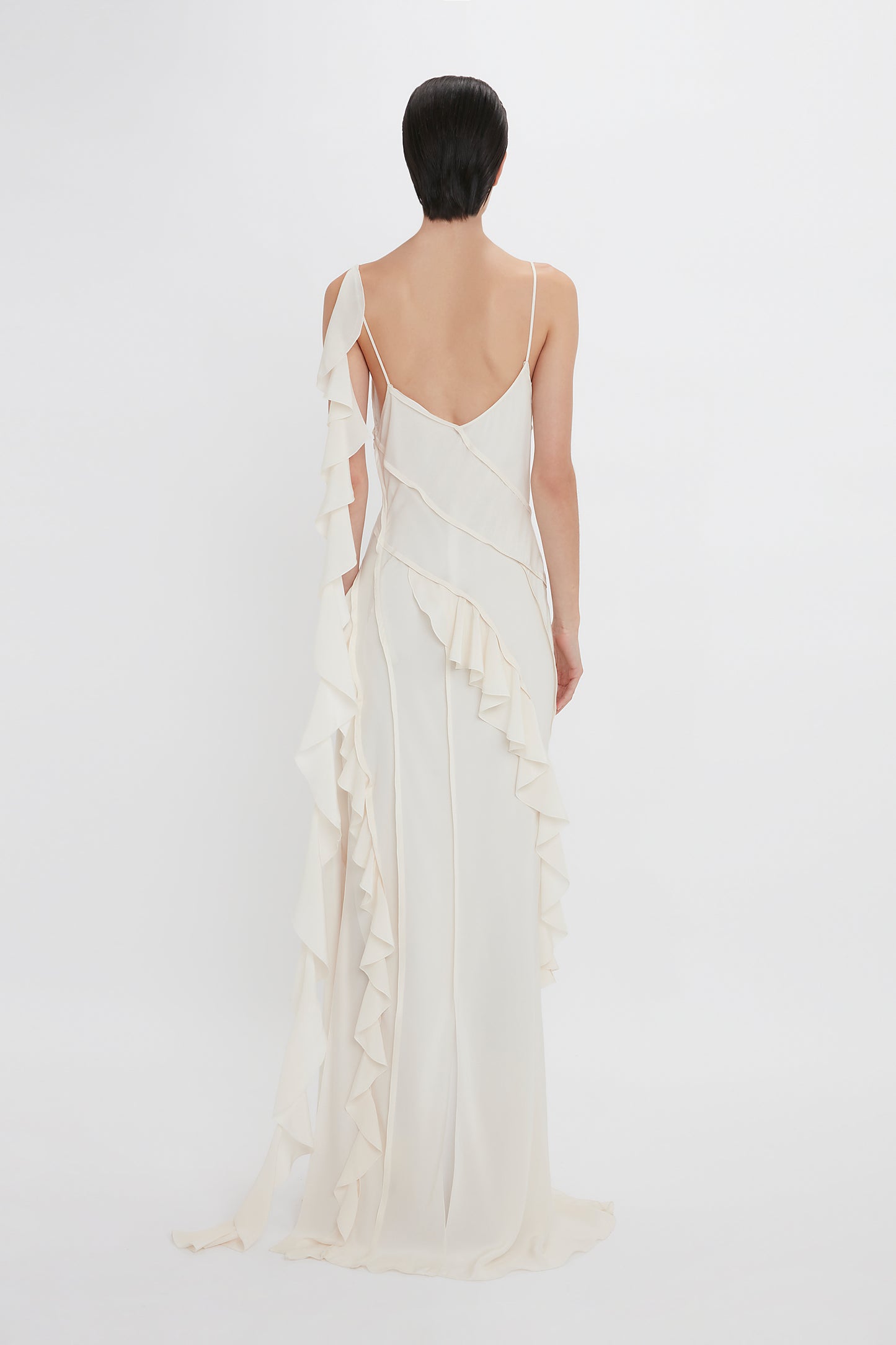 A woman viewed from behind wearing an elegant white Victoria Beckham Exclusive Asymmetric Bias Frill Dress In Ivory with spaghetti straps, against a plain white background.