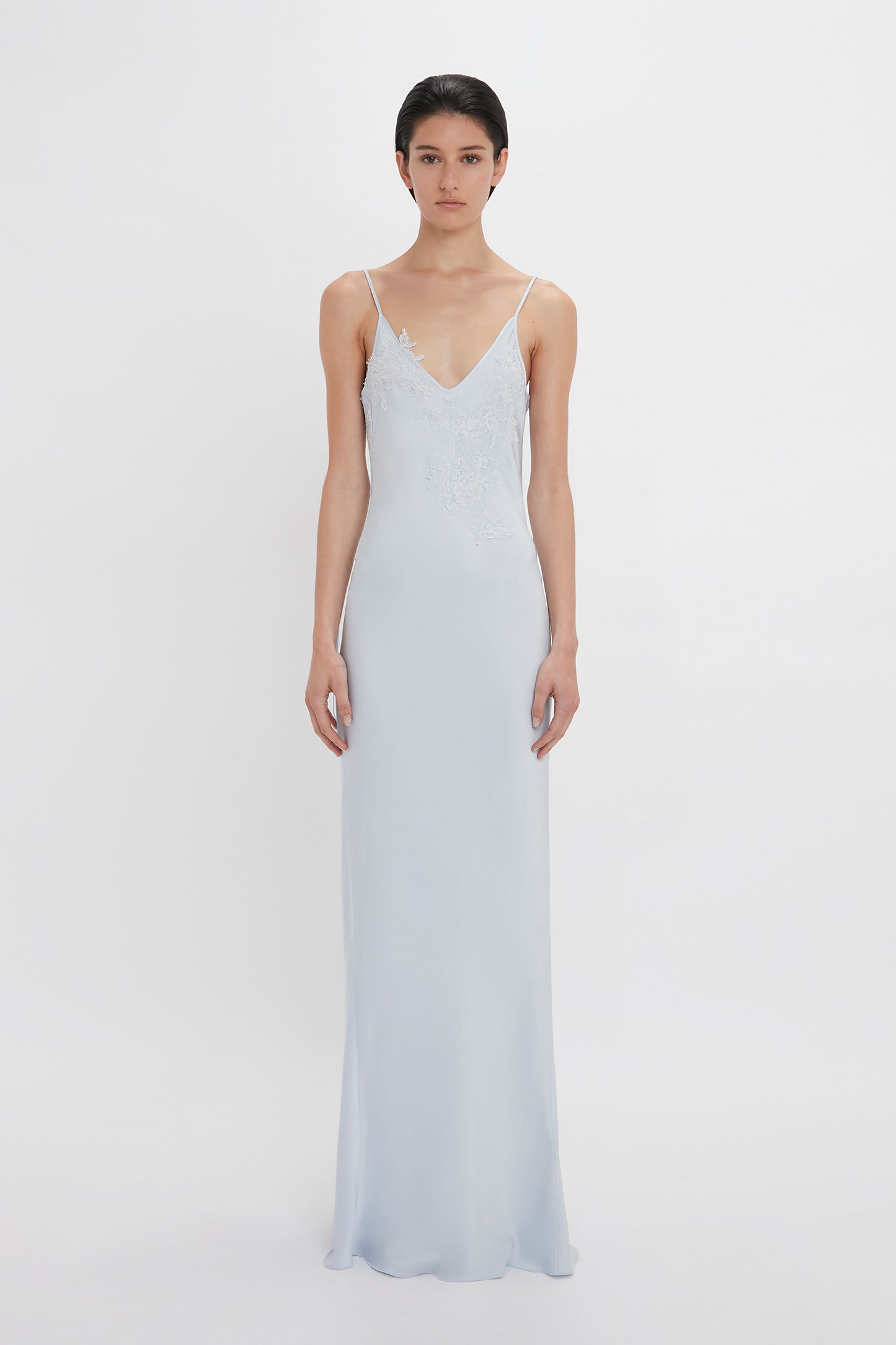 A woman in an Exclusive Lace Detail Floor-Length Cami Dress In Ice by Victoria Beckham, with spaghetti straps and lace detailing at the bodice, standing against a white background.