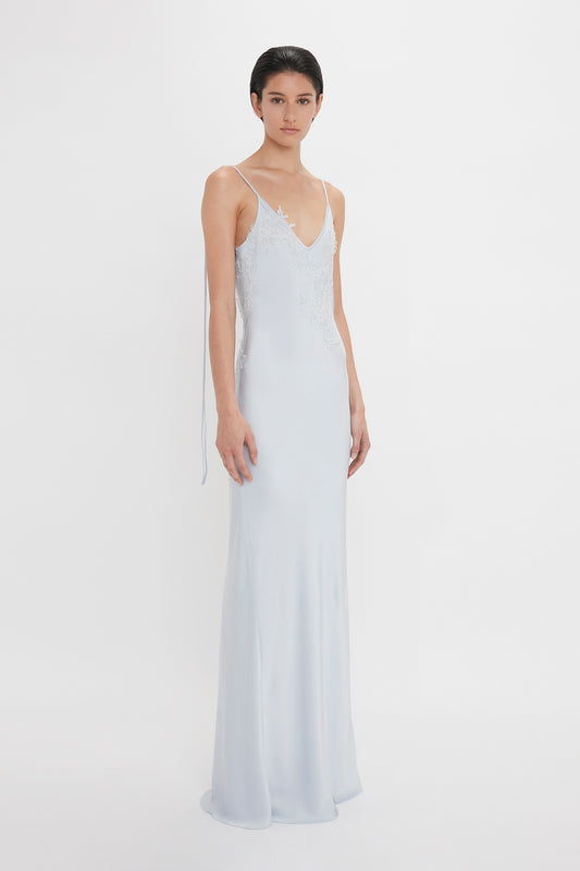 A woman stands against a white background wearing a Victoria Beckham Exclusive Lace Detail Floor-Length Cami Dress In Ice with delicate lace details on the spaghetti straps.