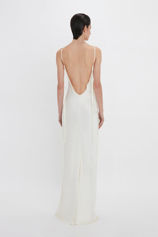 A woman in a Victoria Beckham Exclusive Lace Detail Floor-Length Cami Dress In Ivory, standing against a plain white background, viewed from the back.