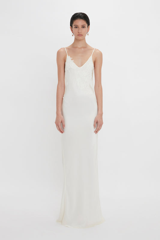 A woman in an elegant white bridal gown with crepe back satin, and lace detailing on the bodice, camisole-style straps, and a flowing skirt, stands against a plain white background. This stunning Exclusive Lace Detail Floor-Length Cami Dress In Ivory by Victoria Beckham is truly breathtaking.